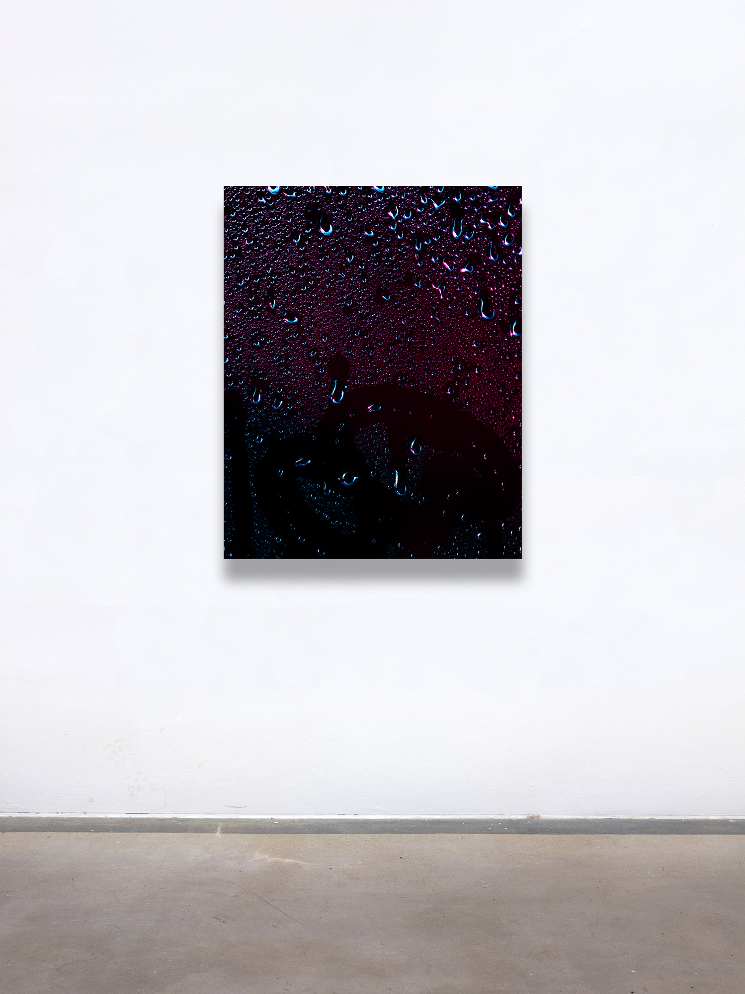 A graduate artwork by Joel Dixon, presented with The Graduate Gallery. The piece features a dark, textured surface adorned with glistening water droplets, set against deep hues of black, purple, and red. The abstract composition evokes a moody, atmospheric tone, with the droplets adding a sense of depth and tactile contrast. Displayed on a minimal white gallery wall, the work draws attention to its intricate details and reflective surface.