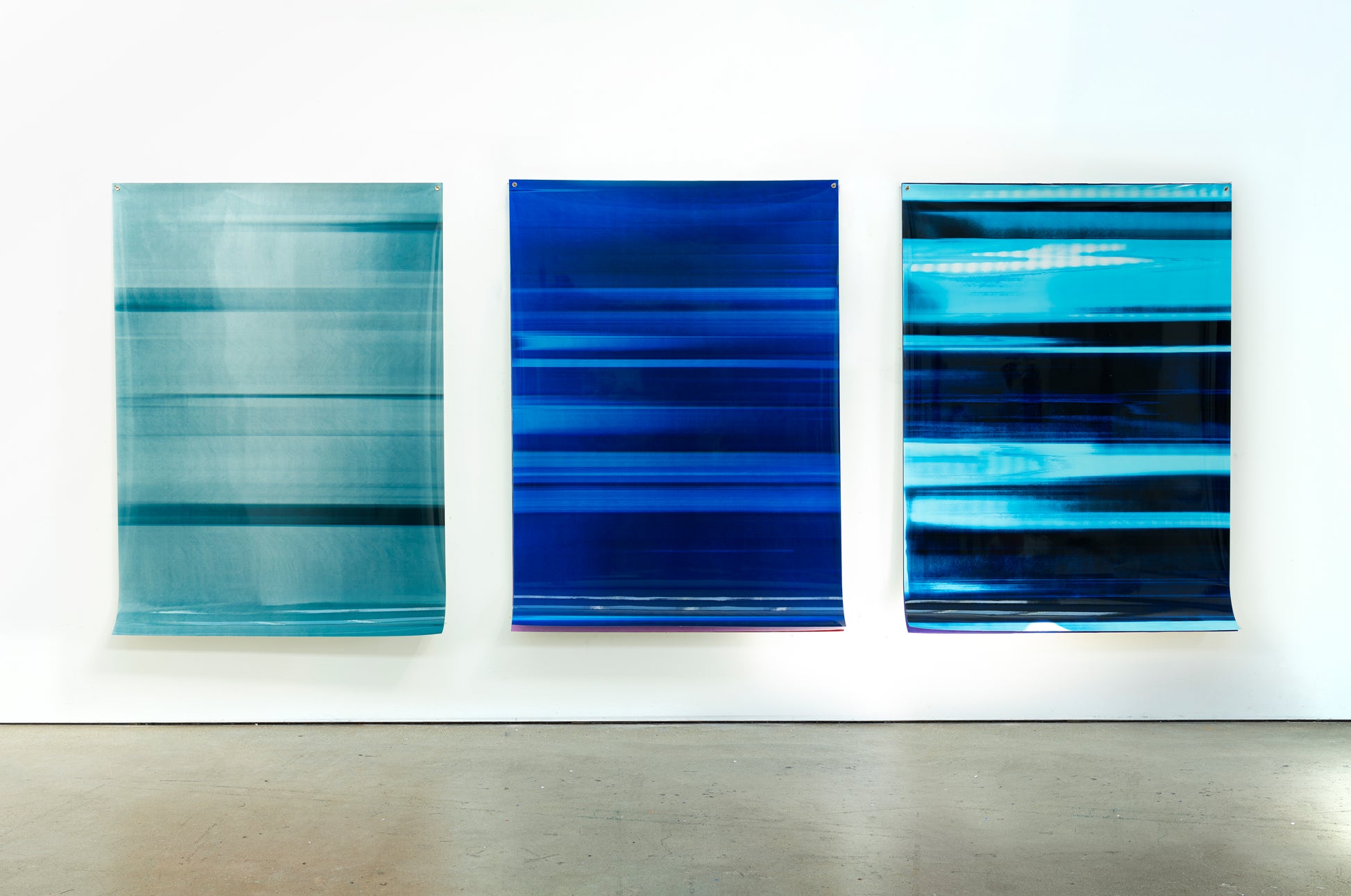 Three abstract artworks by a graduate artist in the UK hang side by side on a white wall. The pieces, available for sale at The Graduate Gallery, feature horizontal stripes in shades of blue. Each artwork has a glossy finish and appears slightly reflective.