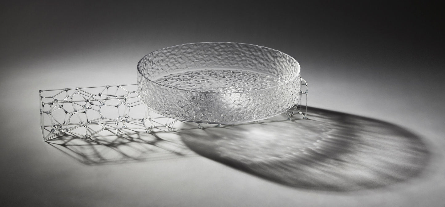 A circular, translucent gray glasswork bowl, crafted by a graduate artist in the UK, rests elegantly on a geometric wireframe base. This intricate network of lines and nodes casts delicate shadows below, reflecting the innovative artworks for sale at The Graduate Gallery.