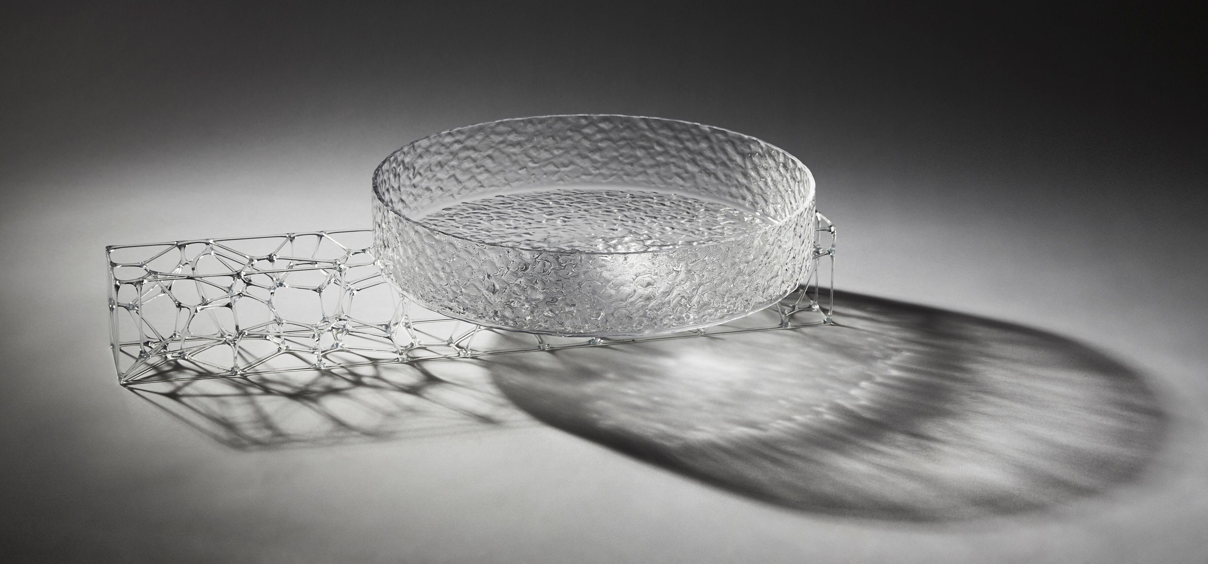 A circular, translucent gray glasswork bowl, crafted by a graduate artist in the UK, rests elegantly on a geometric wireframe base. This intricate network of lines and nodes casts delicate shadows below, reflecting the innovative artworks for sale at The Graduate Gallery.