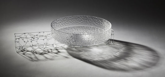 A circular, translucent gray glasswork bowl, crafted by a graduate artist in the UK, rests elegantly on a geometric wireframe base. This intricate network of lines and nodes casts delicate shadows below, reflecting the innovative artworks for sale at The Graduate Gallery.