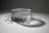 A circular, translucent gray glasswork bowl, crafted by a graduate artist in the UK, rests elegantly on a geometric wireframe base. This intricate network of lines and nodes casts delicate shadows below, reflecting the innovative artworks for sale at The Graduate Gallery.