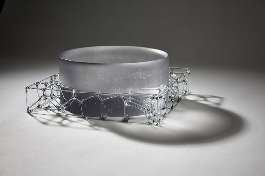 A circular, translucent gray glasswork bowl, crafted by a graduate artist in the UK, rests elegantly on a geometric wireframe base. This intricate network of lines and nodes casts delicate shadows below, reflecting the innovative artworks for sale at The Graduate Gallery.
