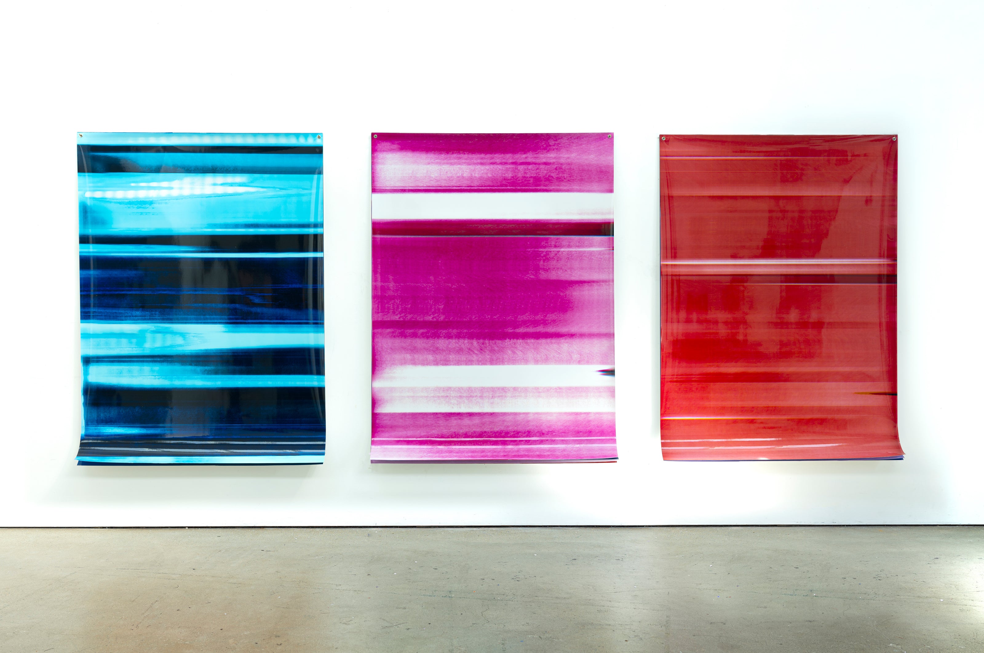 Three abstract artworks by a graduate artist in the UK hang side by side on a white wall. The pieces, available for sale at The Graduate Gallery, feature horizontal stripes in shades of blue, pink, and red. Each artwork has a glossy finish and appears slightly reflective.